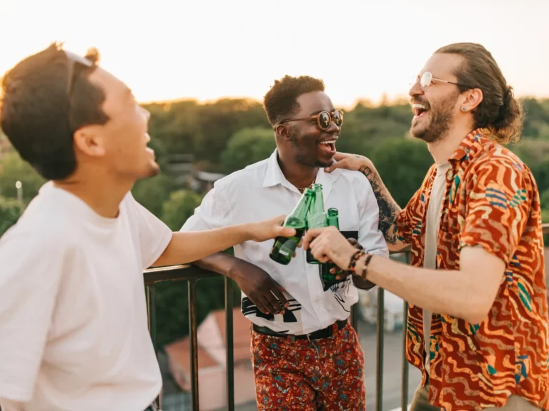Three cheerful friends laughing and toasting with green beer bottles on a sunlit balcony at Two Travel Home.