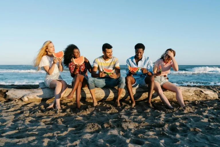 Five happy friends sitting on a wooden log eating watermelon slices on a sandy beach at Two Travel Home.