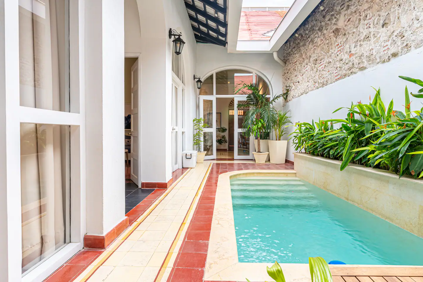 A narrow courtyard at Casa del Torno Cartagena features a small rectangular pool, bordered by potted plants and greenery, alongside white walls and arched windows, with a glass door leading inside.